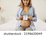 Small photo of Happy mom-to-be thinking of name for her baby. Smiling pregnant lady trying to guess gender of unborn baby. Beautiful young woman sitting on bed with question mark card in hands. Gender reveal concept