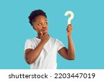 Small photo of Young dark skinned woman with doubtful expression is looking at wooden question mark in her hand on light blue background. Concept of plans, thoughts, ideas for decision making, questions and answers.