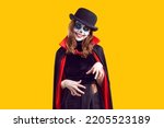Small photo of Preteen girl with creative Halloween make-up and wearing dracula cape isolated on orange background. Portrait of smiling child having fun at carnival party. Halloween party concept. Web banner.