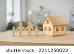 Housing for families. Close up of wooden figurines of family with children and model of small one-story house. Miniature figurines made of light wood stand on table in bright room. Blurred background.