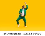Happy young guy having fun at crazy St Patrick's Day holiday party. Full body shot of funny, cheerful, goofy man wearing green suit and sunglasses dancing isolated on bright yellow colour background