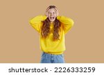 Small photo of Negative human emotions. Emotional teenage girl because of anger and resentment screams and cries on beige background. Girl grabs her head with her hands. Concept of hatred, rage and frustration.