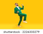 Small photo of Young man celebrating success. Happy funny joyful excited guy in stylish green party suit and cool glasses raising fist up and dancing isolated on bright yellow background. Full length shot, side view