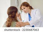 Small photo of Medicine and dermatology. Professional dermatologist examines face of teenage girl with magnifying glass in her office. Young serious female doctor determines condition of skin of teenager's face.