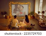Romantic couple having Valentine's Day date at home, using modern projector in living room interior with beautiful fairy lights, watching movie, looking at digital photos, enjoying memories. Back view