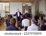 Business trainer in suit and shirt is greeting group of adult business people students in modern office spreading his arms asides. People sitting at desks backs to camera. Adult education concept.