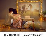 Back view happy loving couple having cozy date at home on Valentine's Day hugging on rug in living room with beautiful lights, watching movie on modern projector, looking at photos, recalling memories