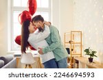Happy young couple hugging and kissing at home. Husband and wife hugging enjoying tender romantic moment against background of red heart shaped balloons and glowing lights