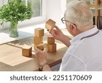 Small photo of Elderly man collects wooden cubes at table opposite the window in modern rehabilitation center for the feeble-minded. Creative idea of training for dementia, feeble-minded, Alzheimer's disease.