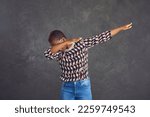 Beautiful young black woman in trendy jumper dancing dub against stone street wall. Happy funky African lady in geometric pattern outfit doing funny dab dance arm move isolated on dark grey background