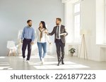 Small photo of People and real estate. Young married couple in love together inspects house shown to them by male realtor. Real estate agent talks about benefits of house to family who walks by him holding hands.