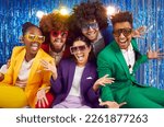 Small photo of Portrait of funny group of happy young people dressed in bright costumes, voluminous curly wigs and colorful large glasses. Group of friends fooling around on the background of blue shiny wall.