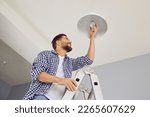 Happy man changing a light bulb at home. High angle shot of a young man in a checkered shirt standing on a step ladder and placing a modern energy-saving lightbulb in a white lamp on the ceiling