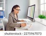 Small photo of Business accountant working in office. Serious busy woman in suit and glasses sitting in front of computer screen, checking book of financial records and using spreadsheet files on PC