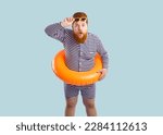Small photo of Funny surprised fat man in beach floatie standing isolated on blue background. Plump man in swimsuit and orange swim ring takes off sunglasses, looks at camera and says WOW. Summer holiday concept