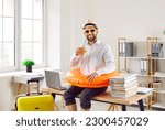 Funny office worker ready to go on summer holiday. Happy smiling man in white shirt, sun hat beach ring and sunglasses drinks orange juice, sitting on desk with laptop and papers. Annual leave concept