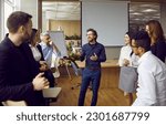 Small photo of Young man business coach trainer talking to group of people in office standing around him and listening. Office workers, entrepreneurs on corporative training, team building seminar or master class.