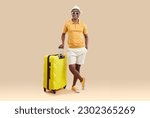 Small photo of Portrait of happy african american traveler with yellow suitcase on light beige background. Full length of smiling millennial man in stylish summer clothes holding handle of suitcase looking at camera