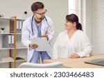 Small photo of Overweight woman having consultation at doctor's office. Portrait of smiling doctor holding report file with appointment and giving consultation to a fat patient during medical examination in clinic.