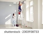 Small photo of Young pretty woman homeowner watching the professional electrician man or technician with screwdriver maintaining, cleaning, repairing or installing modern air conditioner indoors standing on ladder.