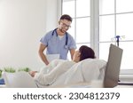 Small photo of Friendly male nurse and female patient at clinic. Smiling young man in scrubs uniform sets intravenous IV line infusion system for overweight woman lying on medical bed in hospital room