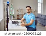 Small photo of Portrait of happy, smiling, young man patient receiving intravenous vitamin infusion through sterile IV line in his arm while sitting at desk with laptop computer at home. Vitamin therapy concept