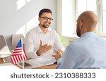 Small photo of Happy, smiling, friendly young male Consul of United States of America sitting at office table with American flag and talking to man about his immigration plans and USA travel visa application