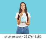 Small photo of Happy smiling brunette young girl in white t-shirt and jeans saving money putting coin in blue piggy bank on blue background looking at camera. Save money, self finance, savings capital concept.