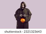 Small photo of Man in grim reaper costume with creepy facial expression collects candy during Halloween celebration. Man in black robe and skeleton makeup holds candy bucket in form of pumpkin on lilac background.
