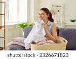 Woman smelling clean laundry. Happy beautiful young housewife sitting on couch with laundry basket, holding perfectly clean, washed, white shirt, smelling fresh, natural aroma, and smiling