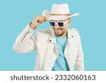 Small photo of Funny Caucasian man in cool sunglasses puts on cowboy hat and looks forward smiling playing toreador or going to trendy themed party dressed in white leather jacket stands on turquoise background