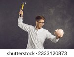 Small photo of Financial difficulties. Emotional and crazy frustrated young man is going to break piggy bank with hammer. Angry man shouting holding piggy bank and hammer in his hands standing on gray background.