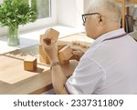 Small photo of Demented senior patient playing brain game. Old man with Alzheimer's disease looking at wooden cube that he's holding in hand while sitting at desk in geriatric clinic