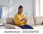 Small photo of Worried young woman looking st cell phone screen while sitting on couch at home. Confused upset attractive African American girl got unpleasant news in message, thinking over received notification