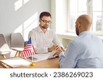 Small photo of Serious strict male worker at United States of America Consulate sitting at office table with American flag looking at filled out visa application form of young man. USA travel and immigration concept