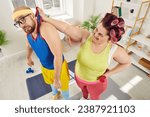 Small photo of Funny dominant female fat woman leader raising her boyfriend in sportswear by hand dominating him during doing sport exercises with dumbbells at home. Domination, submission and violence concept.