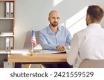 Small photo of Consul at Consulate of United States of America sitting at office table with American flag and listening to young man telling about his problems with immigration and USA travel visa application