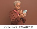 Small photo of Young dazed ethnic African American woman holds phone and covers open mouth with hand indignant at news or inaccurate statement on Internet site stands on brown background. Surprise, indignation