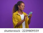 Small photo of Young optimistic long haired African American woman using mobile phone with slight smile dialing friend number wants to make call to gossip or arrange date stands on lilac studio background