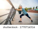 Small photo of A senior woman stretches during her workout. Mature woman exercising. Portrait of fit elderly woman doing stretching exercise in park. Senior sportswoman making stretch exercises