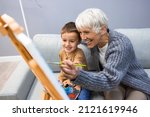 Senior woman with child painting on canvas. Grandmother spending happy time with grandson. Small boy listening to his grandmother who is teaching him to paint on canvas at home.