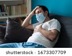 A sick caucasian Man wearing face mask feeling sick headache and cough because of Coronavirus Covid-19 on the sofa in quarantine
