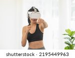 Small photo of Athletic Asian young woman in sportswear using VR virtual reality glasses headset workout boxing excercise training at home.Healthy female punching boxing footwork cardio game exercise in metaverse