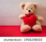 Valentines day. Teddy bear holding a big heart sitting on red floor, wooden wall background, copy space, valentines card template