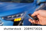 Small photo of Man uses thumb to press black button on remote control key removing alarm signalling from bright blue car and headlights flash closeup