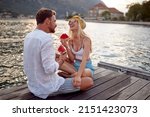 Couple having good time on date by sea siting on wooden dock, laughing and eating watermelon. Love, fun, togetherness concept.