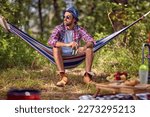 Young handsome hipster male sitting in hammock holding beer. Picnic in forest. Holiday, leisure, lifestyle concept.