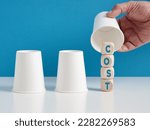 Finding the hidden costs concept. Man playing the shell game with three cups reveals the word cost on wooden cubes.