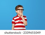 Nerdy boy in eyeglasses and red striped shirt rubbing chin and looking away while thinking near empty space against blue background