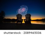 Small photo of Canada day fireworks on a lake in Muskoka, Ontario Canada. On the wooden dock two Adirondack chairs are facing the sunset orange hues
